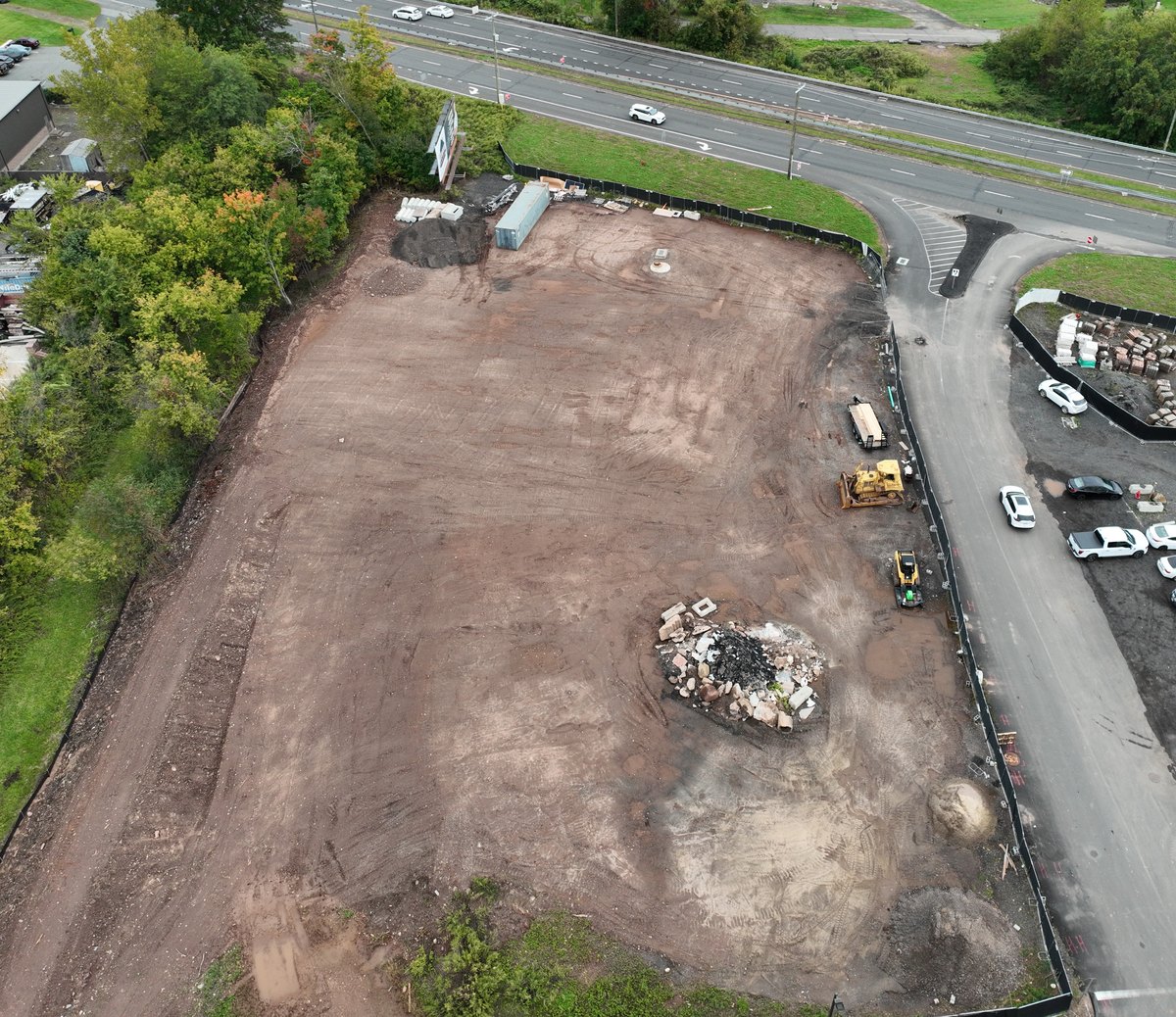 Aerial oblique of a cleared development site before vertical construction, with the surrounding commercial context