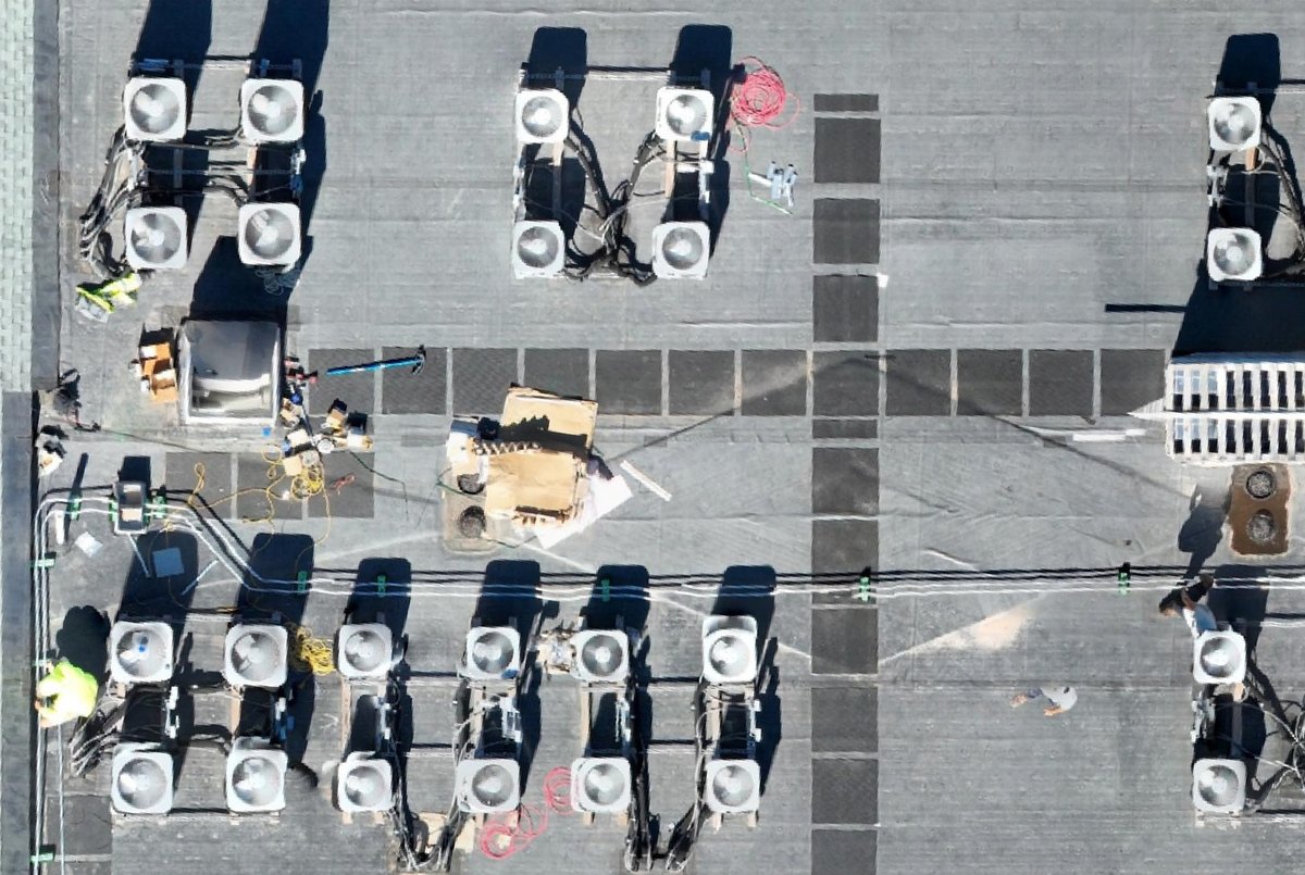 Close orthomosaic of a rooftop mechanical zone: banks of condensing units on sleepers, refrigerant line sets and electrical whips, walkway pads, a roof hatch, and staged material on the deck